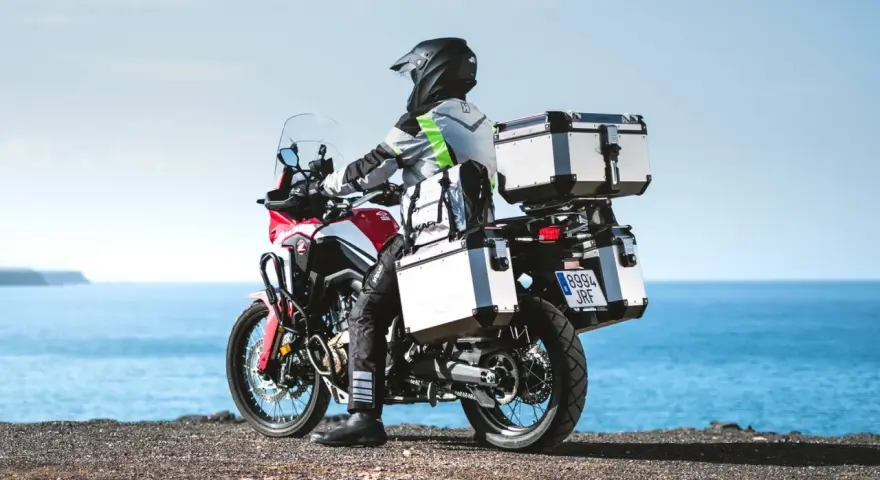 Motociclista con equipaje de **marca Kappa** listo para la aventura junto al mar.