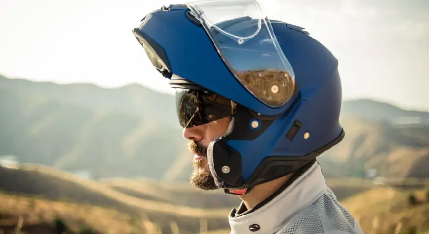Hombre con barba y gafas de sol lleva un casco integral que se abre, azul, con visera levantada, listo para la aventura en la montaña.