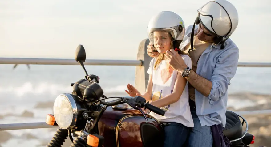 Un hombre ajusta el casco a un niño en una moto, listos para una aventura junto al mar.