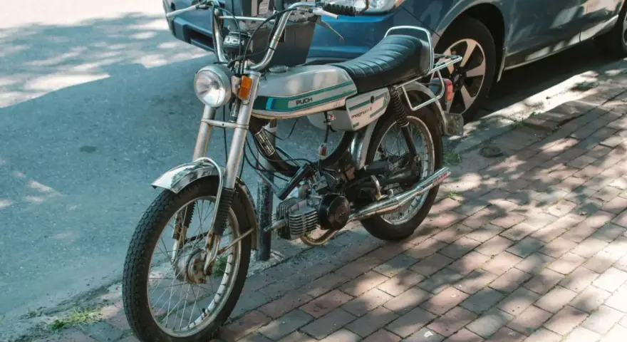 Ciclomotor antiguo Puch Magnum II plateado, listo para matricular. Aparcado junto a un coche azul en la acera.