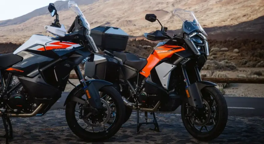 Dos motos KTM Super Adventure listas para la aventura. Modelos naranjas y blancos, con maletas, en un paisaje árido.
