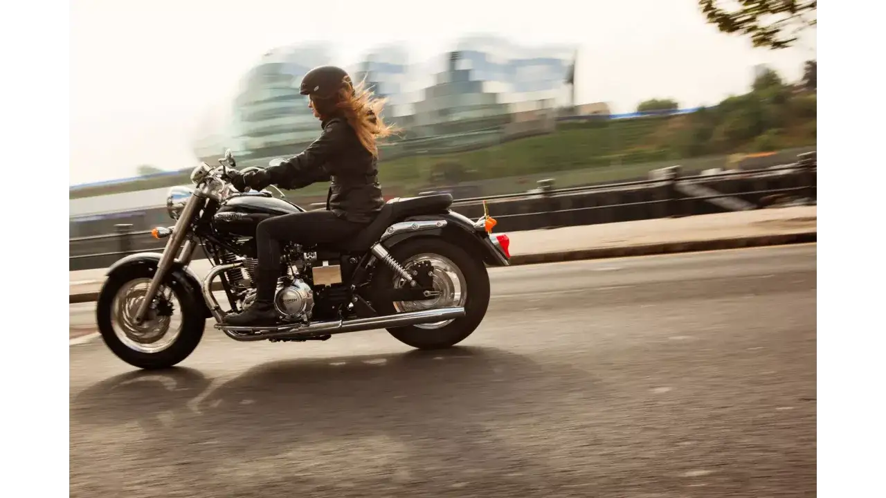Mujer en moto Triumph America 900, con el viento en el pelo, disfrutando de un viaje urbano.