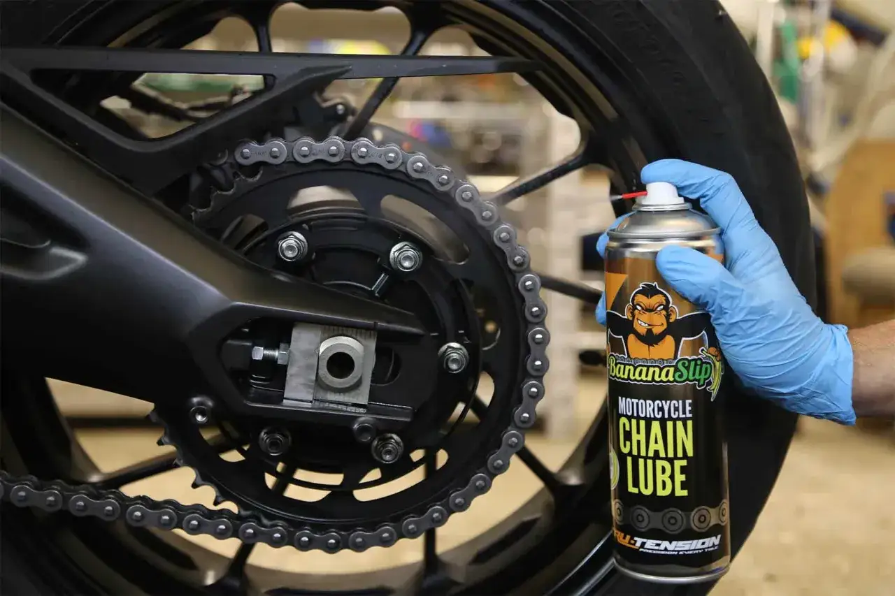 Mano con guante azul aplica lubricante a la cadena de moto. ¡Es hora de engrasar cadena moto para un viaje suave!