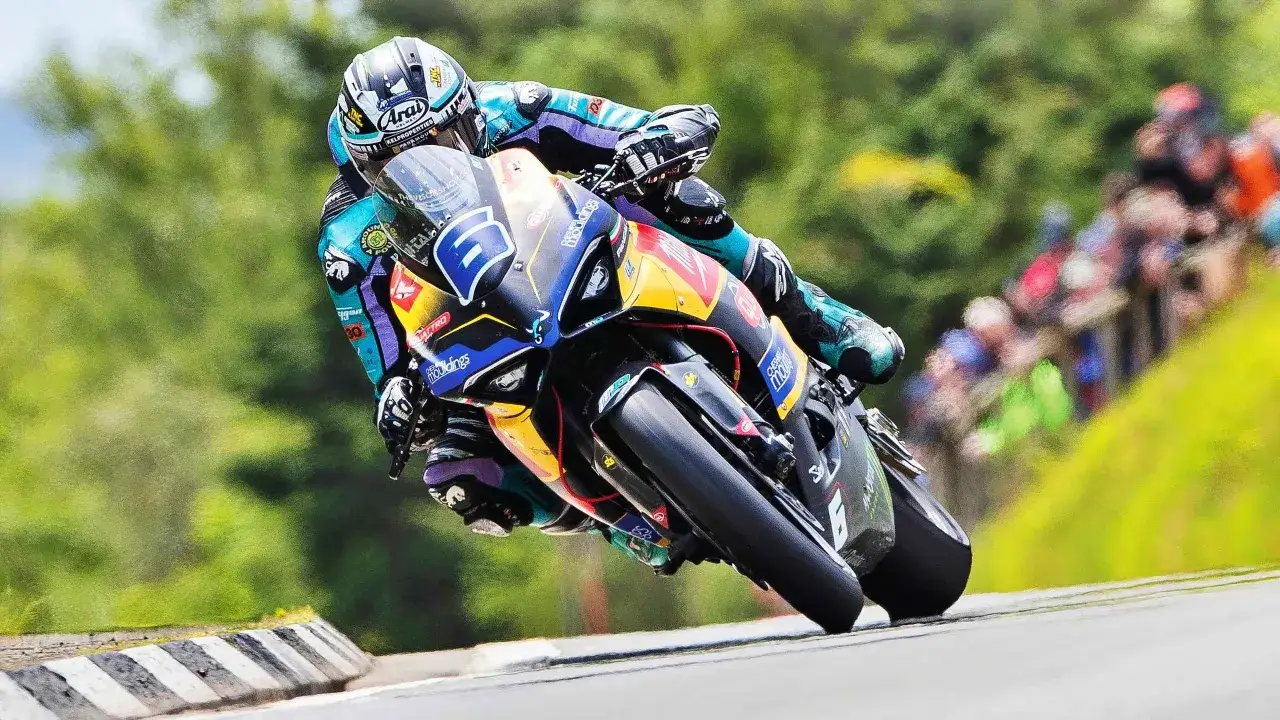 William Dunlop inclinado en su moto de carreras, con el número 6 visible. El fondo verde y la multitud sugieren una carrera emocionante.
