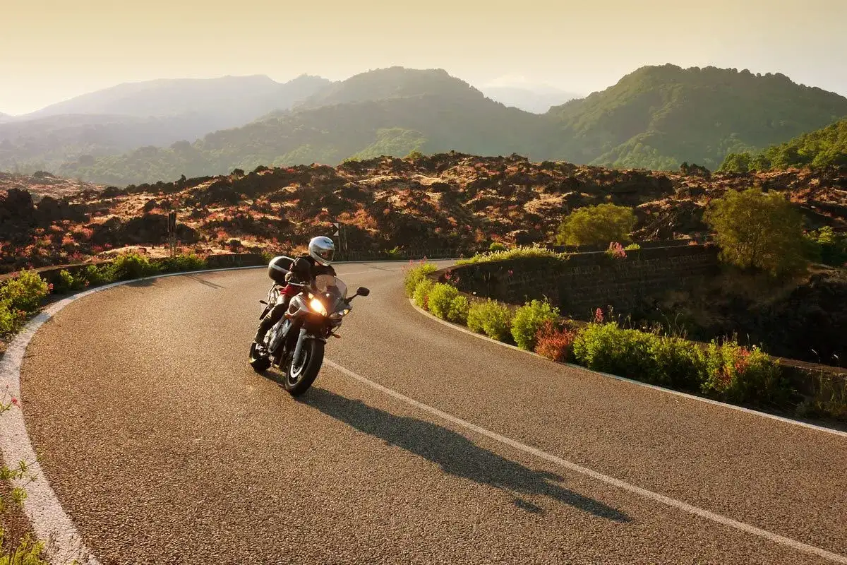 Un motociclista recorre una carretera sinuosa en Cerdeña, rodeado de un paisaje montañoso y vegetación exuberante.