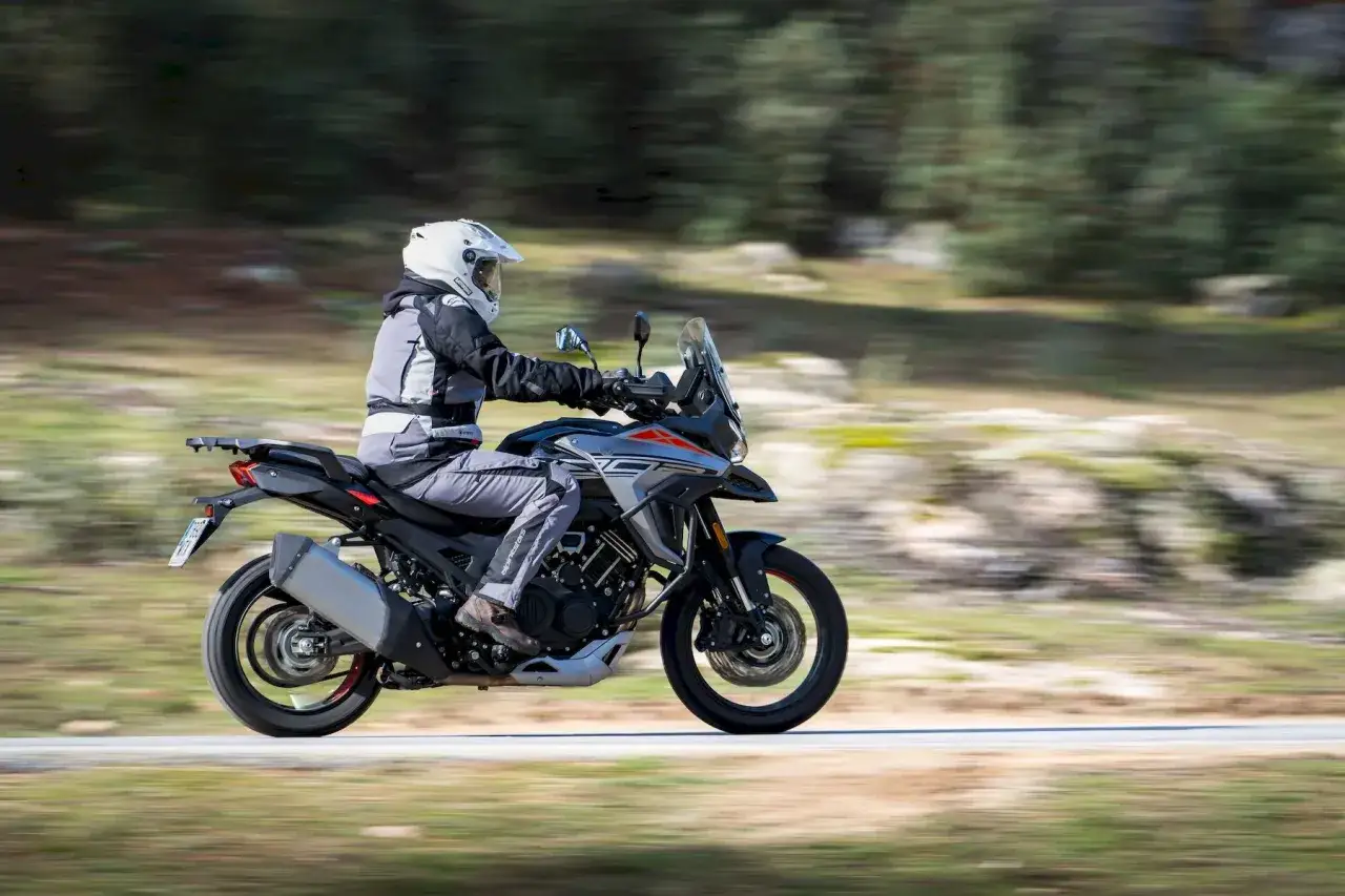 Motociclista con casco blanco y equipo de aventura conduce una moto china trail por un camino rural con &aacute;rboles borrosos al fondo.