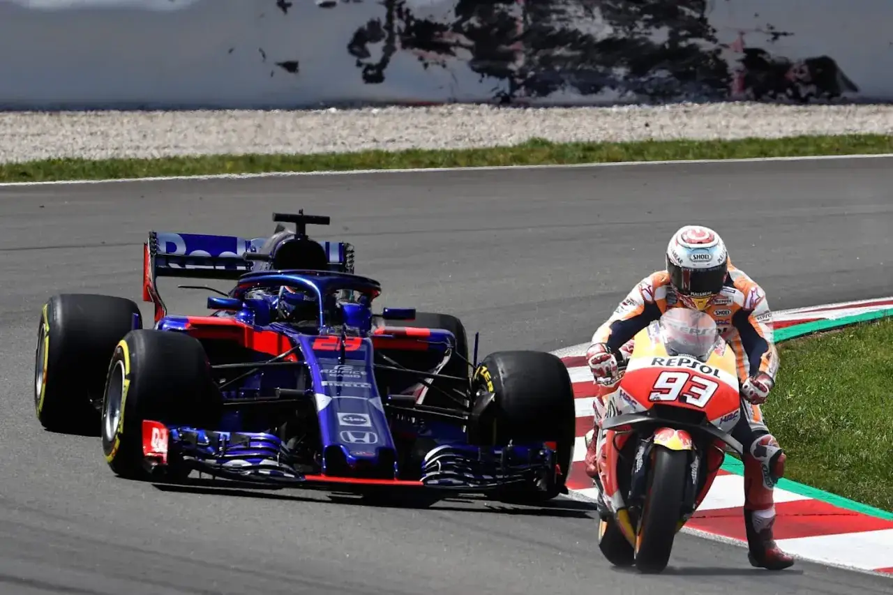 Un coche de F1 y una moto de MotoGP compiten en la pista. El coche azul y rojo avanza rápido, mientras que la moto naranja y blanca con el número 93 se inclina en una curva.