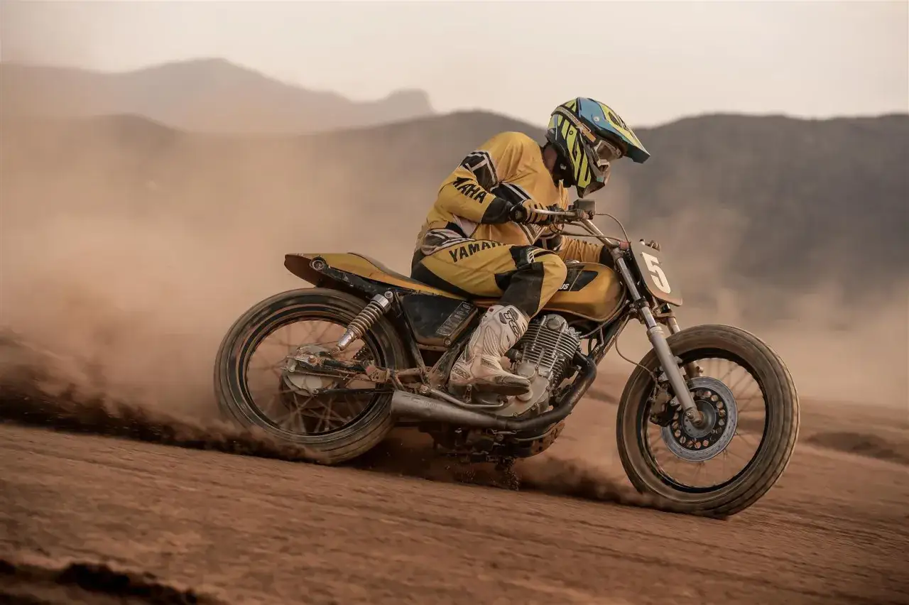 Piloto con casco y traje amarillo compite en una moto por una dirt track, levantando polvo en un paisaje des&eacute;rtico.