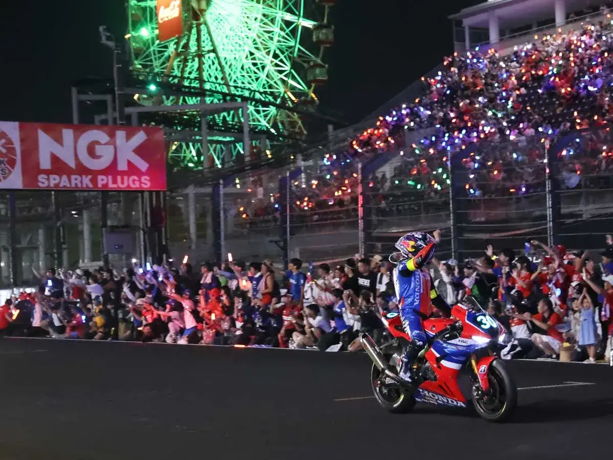 Piloto celebra victoria en las 8 horas de Suzuka. Multitud enciende luces en las gradas, con noria iluminada al fondo.
