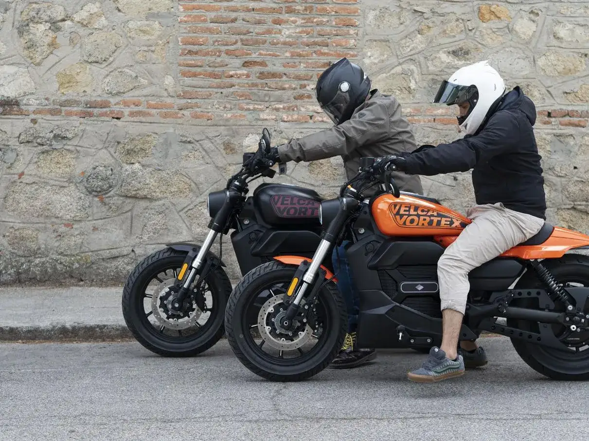 Dos motociclistas con cascos posan junto a dos motos custom, una negra y otra naranja, listas para emprender un viaje. Son las mejores motos custom para viajar.
