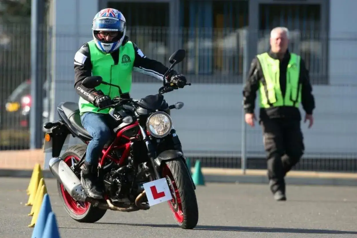 Motociclista con chaleco verde y casco, realizando un examen de circulaci&oacute;n A2. Un instructor con chaleco amarillo observa.