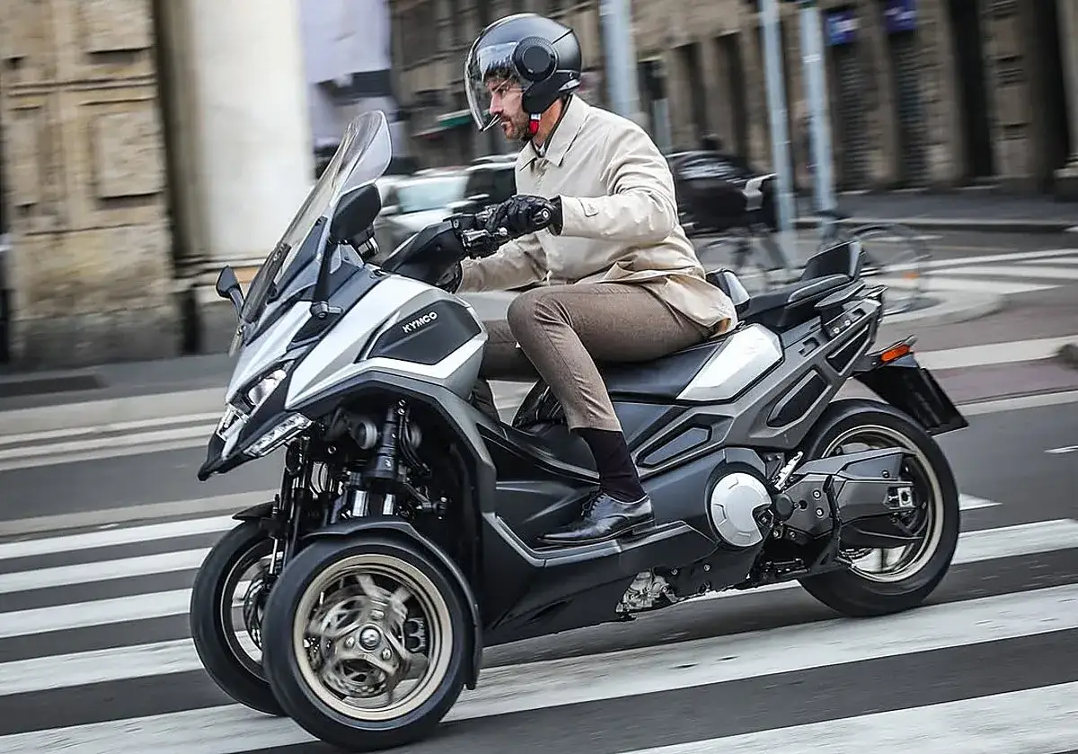 Hombre con casco conduce un triciclo Kymco, ideal para motos para ciudad, cruzando un paso de peatones.