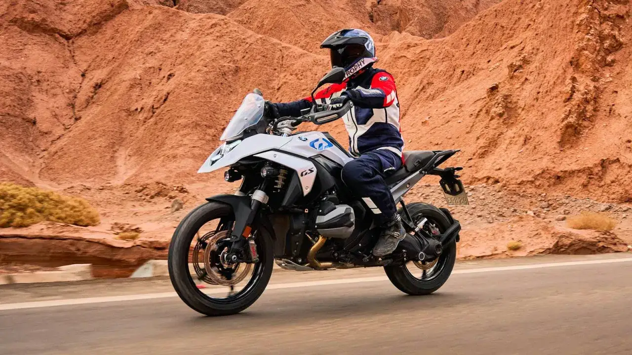 Motociclista en una moto crossover blanca recorre un camino des&eacute;rtico con monta&ntilde;as rojizas de fondo.