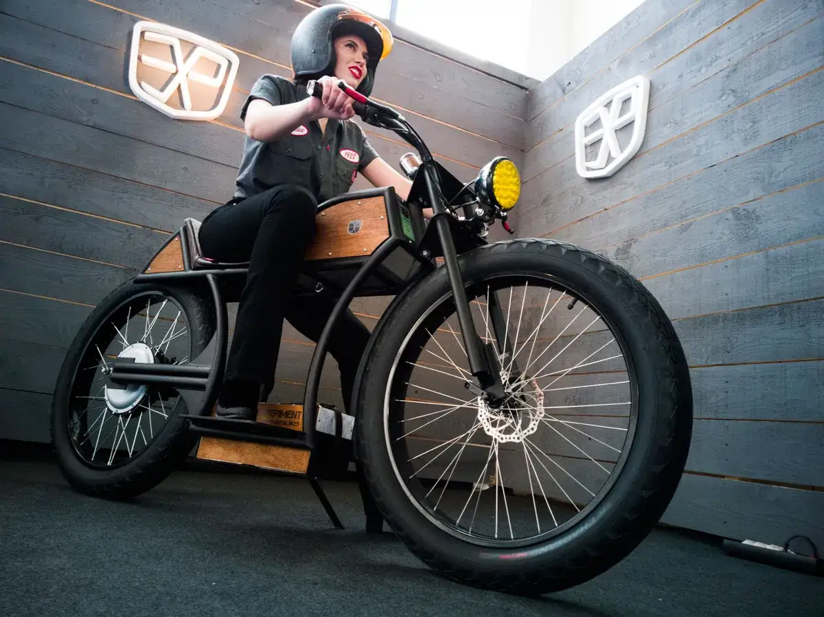 Mujer con casco posa en una moto custom electrica de dise&ntilde;o &uacute;nico, con detalles en madera y rueda delantera ancha.